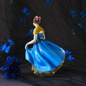 May include: A porcelain figurine of a person with brown hair styled in an updo with a pink bow, wearing a yellow shawl over a vibrant blue dress with a flared skirt. The figure is captured in a dynamic pose, appearing to lift the hem of the dress.