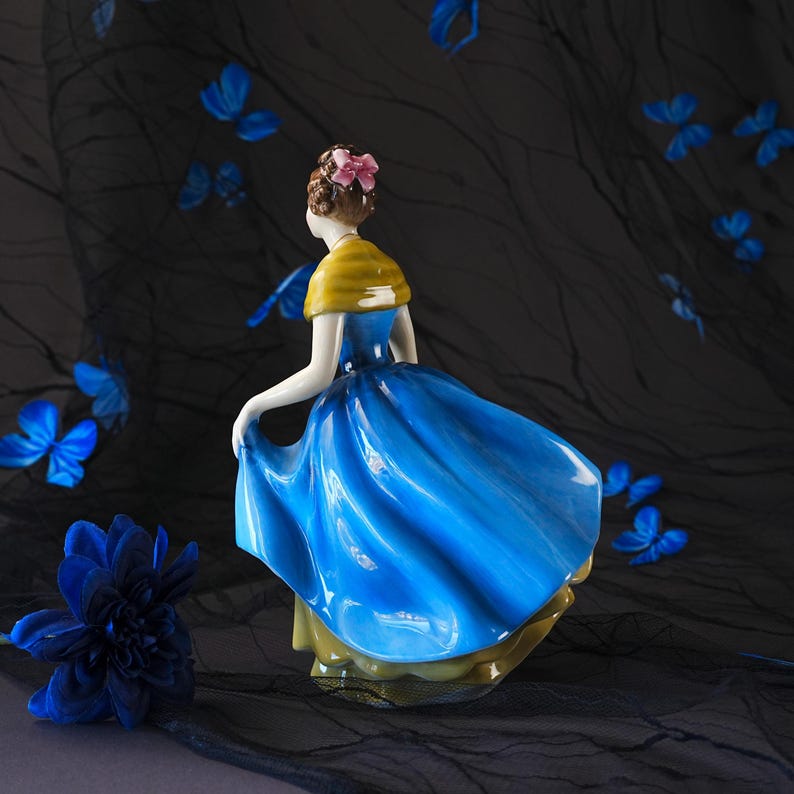 May include: A porcelain figurine of a person with brown hair styled in an updo with a pink bow, wearing a yellow shawl over a vibrant blue dress with a flared skirt. The figure is captured in a dynamic pose, appearing to lift the hem of the dress.
