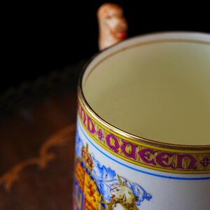 Puede incluir: Una taza de cer&aacute;mica blanca con borde y asa dorados. La taza presenta un dise&ntilde;o colorido con la palabra "Queen" en letras rosas y un escudo de armas. El interior de la taza es de color amarillo claro.