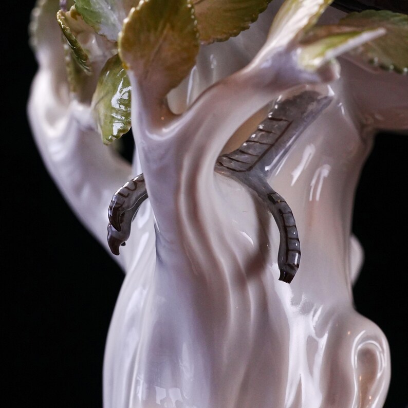 May include: A detailed porcelain figurine featuring a tree with delicate leaves and a bird's claw. The sculpture is primarily white with subtle pink and green accents, showcasing intricate craftsmanship. The claw is a dark grey color.