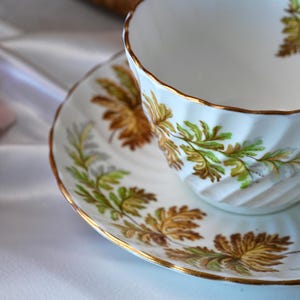 Op de afbeelding: Witte theekop en schotel set met gouden rand en een botanisch ontwerp in groen- en goudtinten. De theekop heeft een gegroefd ontwerp. De schotel en kop hebben een gouden rand.