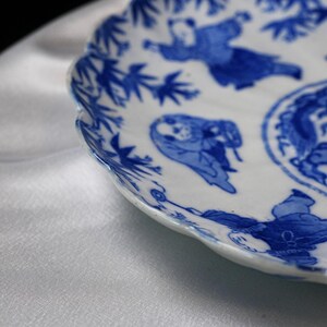 Puede incluir: Plato de cer&aacute;mica blanca con borde festoneado, decorado con ilustraciones azules de figuras y follaje. El plato tiene un dise&ntilde;o asi&aacute;tico tradicional, con ilustraciones detalladas y un acabado brillante.