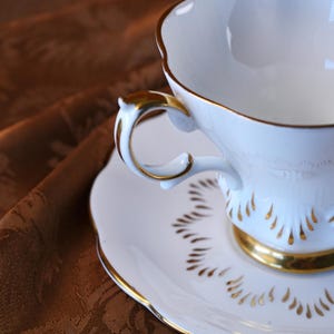 May include: A white teacup and saucer set with gold trim. The teacup has a curved handle and a scalloped rim. The saucer features a matching gold trim and a decorative pattern. The set is designed for serving tea.