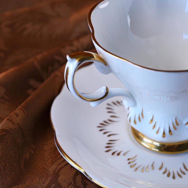 May include: A white teacup and saucer set with gold trim. The teacup has a curved handle and a scalloped rim. The saucer features a matching gold trim and a decorative pattern. The set is designed for serving tea.