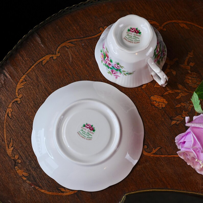 Op de afbeelding: Een witte Royal Albert theekop en schotel met bloemmotief. De kop is ondersteboven en toont de Royal Albert stempel. De schotel heeft een geschulpte rand. De set staat op een houten tafel.