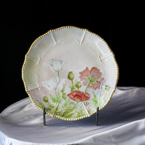 May include: A decorative porcelain plate featuring a hand-painted floral design. The plate has a scalloped edge with a gold trim and a soft pink and white background. The flowers are red and white poppies with green leaves.