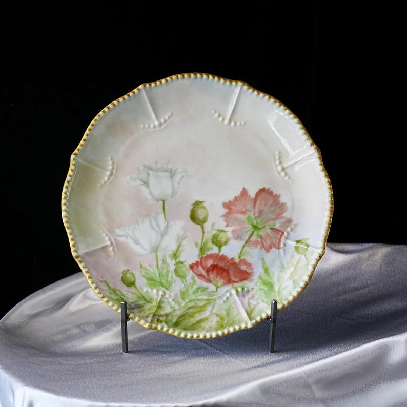 May include: A decorative porcelain plate featuring a hand-painted floral design. The plate has a scalloped edge with a gold trim and a soft pink and white background. The flowers are red and white poppies with green leaves.