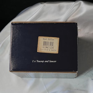 Może przedstawiać: Granatowe pudełko z etykietą z napisem "Royal Doulton AUTUMNS GLORY 2 X TEACUP & SAUCER". Prostokątne pudełko prawdopodobnie zawiera zestaw filiżanki i spodka.