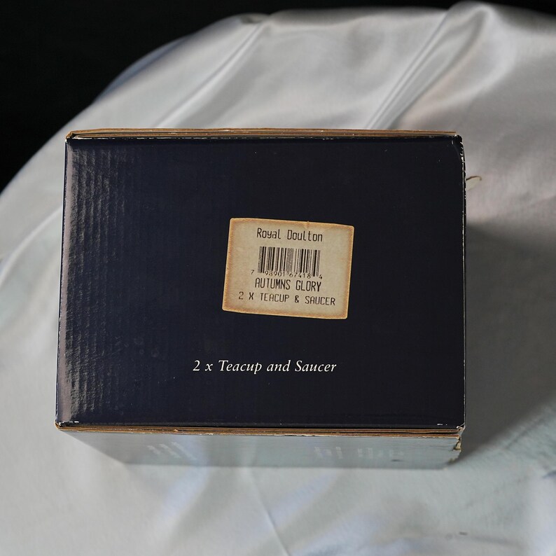 Może przedstawiać: Granatowe pudełko z etykietą z napisem "Royal Doulton AUTUMNS GLORY 2 X TEACUP & SAUCER". Prostokątne pudełko prawdopodobnie zawiera zestaw filiżanki i spodka.