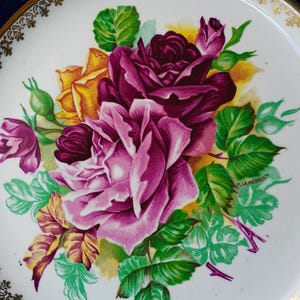 May include: A white ceramic plate featuring a central floral illustration of deep purple, magenta, and yellow roses with green leaves. The rim of the plate is decorated with a gold filigree pattern. The artist signature M. Langbroek is visible.