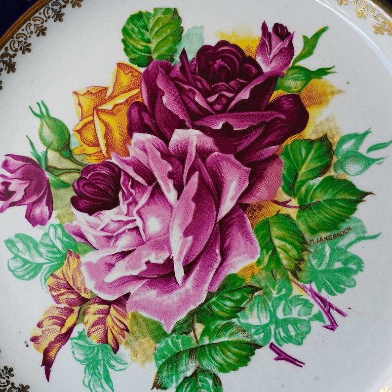 May include: A white ceramic plate featuring a central floral illustration of deep purple, magenta, and yellow roses with green leaves. The rim of the plate is decorated with a gold filigree pattern. The artist signature M. Langbroek is visible.