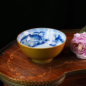 May include: A small, antique bowl with a yellow exterior and a blue and white landscape design on the interior. The bowl sits on a wooden table next to a pink rose. The bowl is a decorative item.