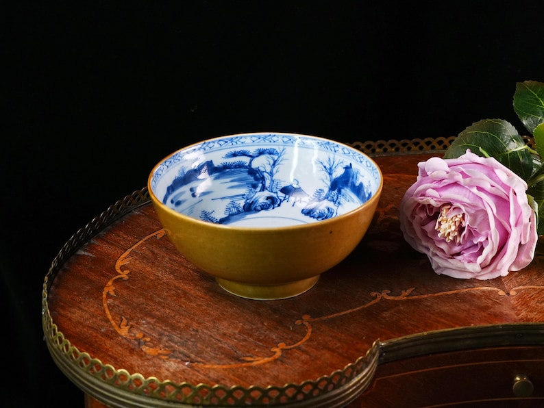 May include: A small, antique bowl with a yellow exterior and a blue and white landscape design on the interior. The bowl sits on a wooden table next to a pink rose. The bowl is a decorative item.