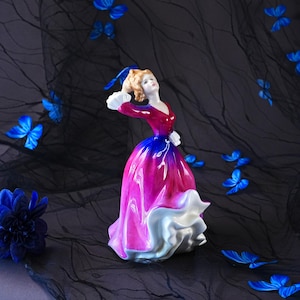 May include: A porcelain figurine of a person in a vibrant, flowing gown that transitions from deep red at the bodice to a bright magenta and blue skirt, finished with a white ruffled hem and white cuffs, standing amidst scattered blue butterflies.