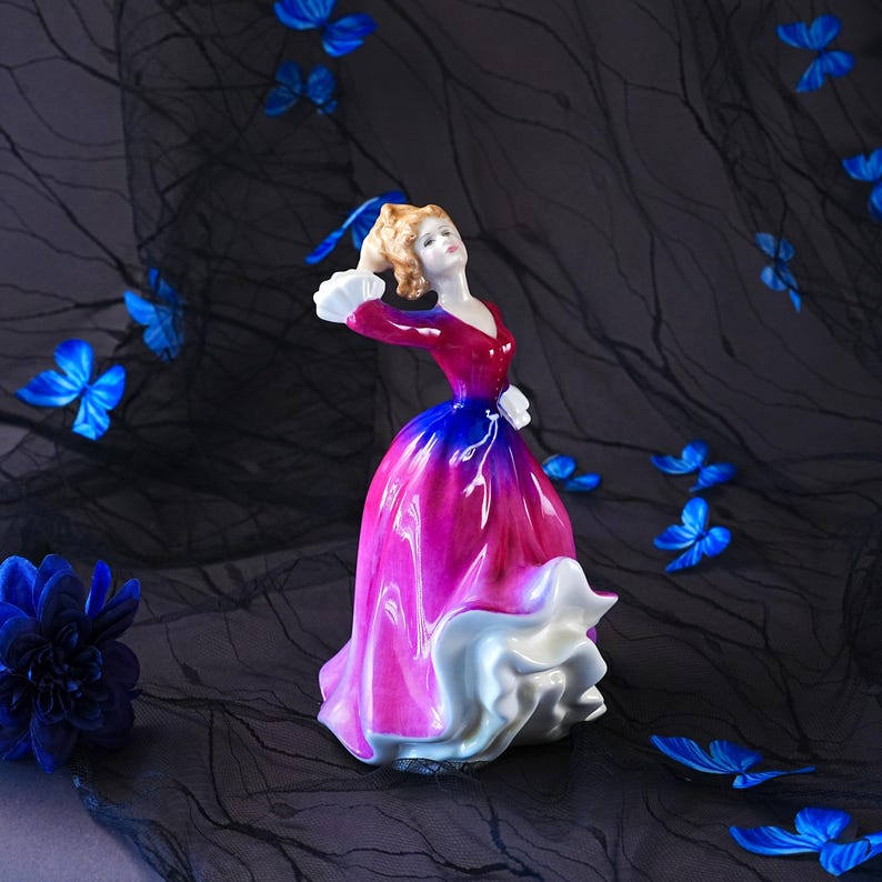 May include: A porcelain figurine of a person in a vibrant, flowing gown that transitions from deep red at the bodice to a bright magenta and blue skirt, finished with a white ruffled hem and white cuffs, standing amidst scattered blue butterflies.