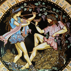 May include: Decorative plate featuring a classical scene with two figures. One plays a flute, the other holds a cup, surrounded by grape vines. The plate has a black background with gold and blue accents.