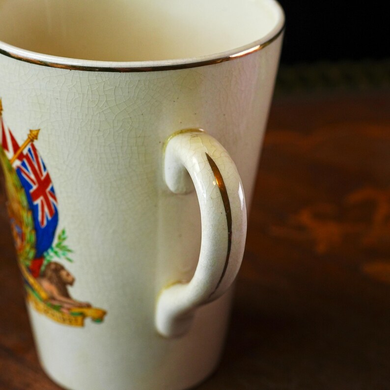 Puede incluir: Taza de cer&aacute;mica color crema con borde y detalles del asa dorados. La taza presenta un emblema detallado con un le&oacute;n, banderas y otros elementos her&aacute;ldicos. El asa es curva y tiene una franja dorada en el centro.