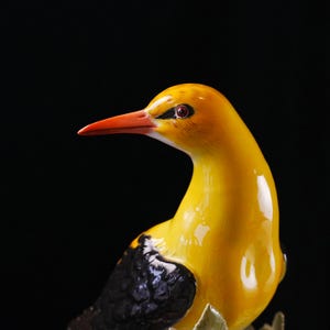 May include: A ceramic bird figurine with a glossy yellow body, a bright orange beak, and black wing details. The bird has a detailed eye and is set against a dark background. The figurine is a decorative item.