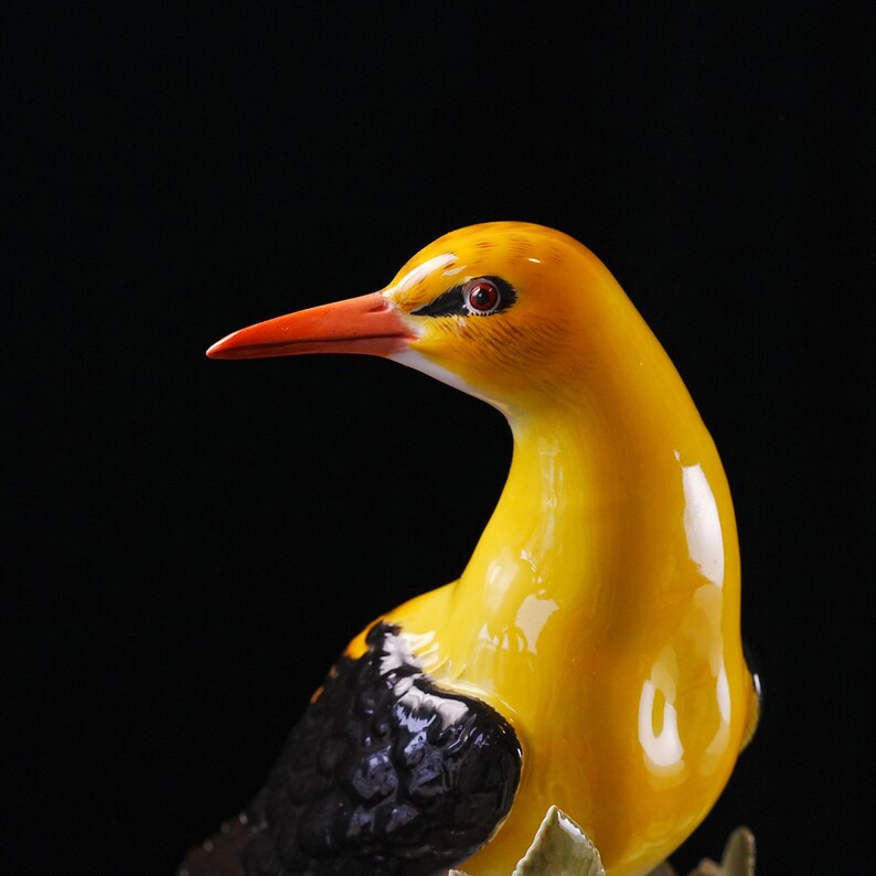 May include: A ceramic bird figurine with a glossy yellow body, a bright orange beak, and black wing details. The bird has a detailed eye and is set against a dark background. The figurine is a decorative item.