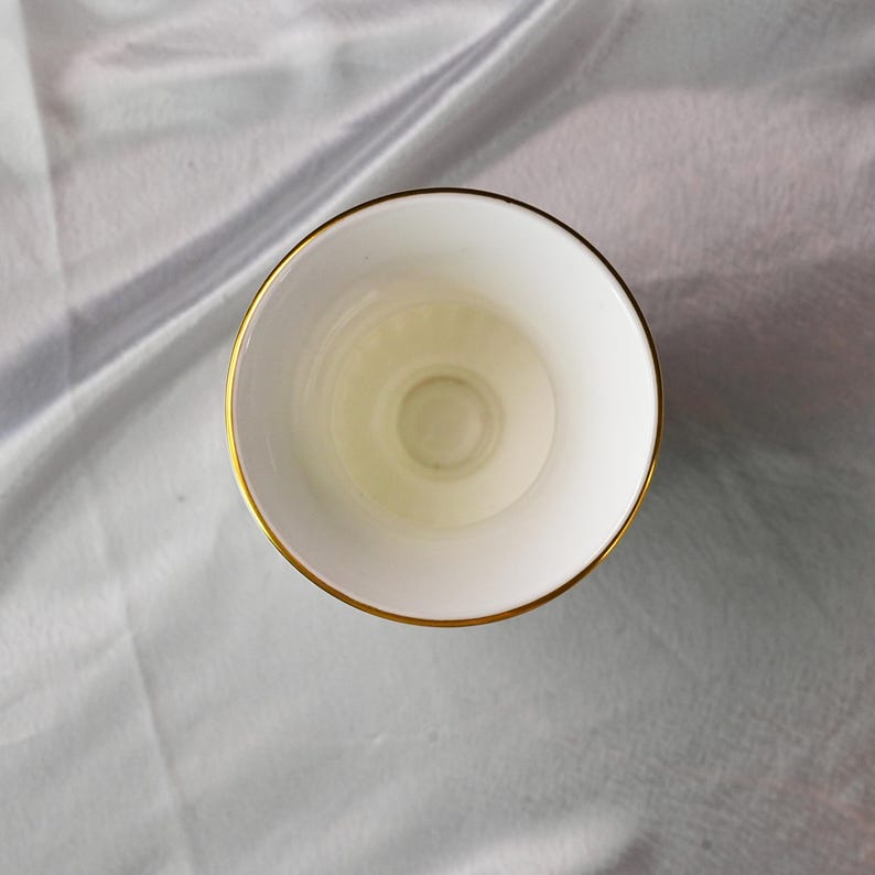 Op de afbeelding: Een witte theekop met een gouden rand, van bovenaf gezien. De binnenkant van de kop is wit met een lichte gele tint. De kop staat op een lichtgrijze, getextureerde achtergrond.