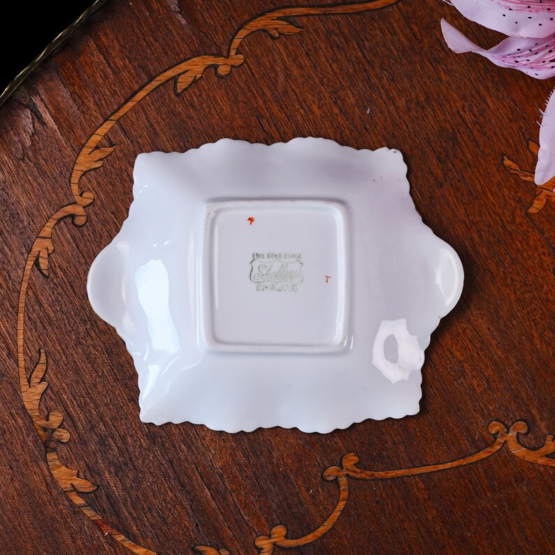 May include: A white square porcelain dish with scalloped edges and two small side handles. The underside features a green stamp with the text Fine Bone China Shelley England, along with small orange paint marks on the white glazed surface.
