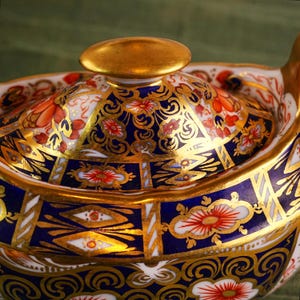 May include: Close-up of a decorative porcelain teapot lid. The lid features a gold knob and is adorned with intricate patterns in navy blue, gold, red, and white. The design includes floral and geometric motifs, showcasing a classic aesthetic.