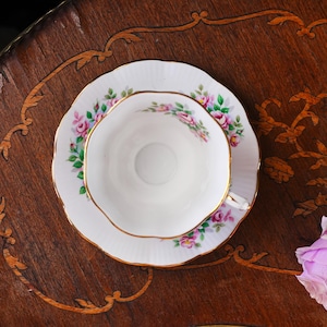 Op de afbeelding: Een witte theekop en schotel met een bloemenrozenpatroon in roze en groen, geaccentueerd met goud. De theekop staat op een bijpassende schotel, beide op een donker houten oppervlak.