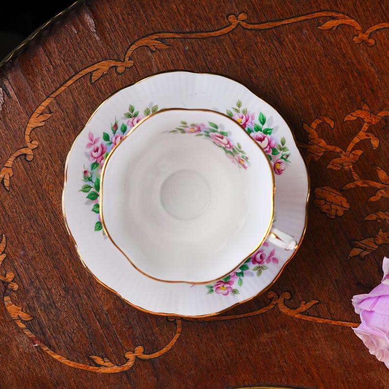 Op de afbeelding: Een witte theekop en schotel met een bloemenrozenpatroon in roze en groen, geaccentueerd met goud. De theekop staat op een bijpassende schotel, beide op een donker houten oppervlak.