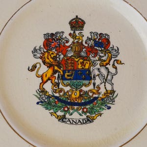 May include: A cream-colored ceramic plate featuring a detailed, colorful coat of arms of Canada in the center. The design includes a lion, a unicorn, a crown, and a banner at the bottom with the text CANADA. The plate surface has a visible crackle glaze.