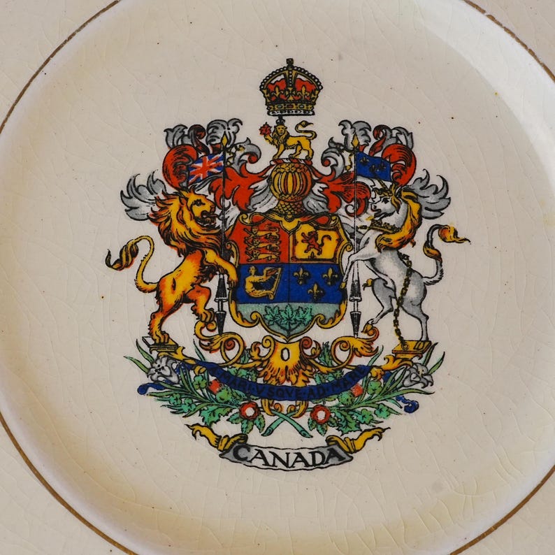 May include: A cream-colored ceramic plate featuring a detailed, colorful coat of arms of Canada in the center. The design includes a lion, a unicorn, a crown, and a banner at the bottom with the text CANADA. The plate surface has a visible crackle glaze.