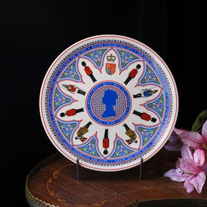 Puede incluir: Un plato conmemorativo de cer&aacute;mica blanca con una silueta azul de la reina Isabel II en el centro, rodeado de ilustraciones de guardias reales, un escudo de armas y motivos florales. El texto dice Silver Jubilee 1952-1977.