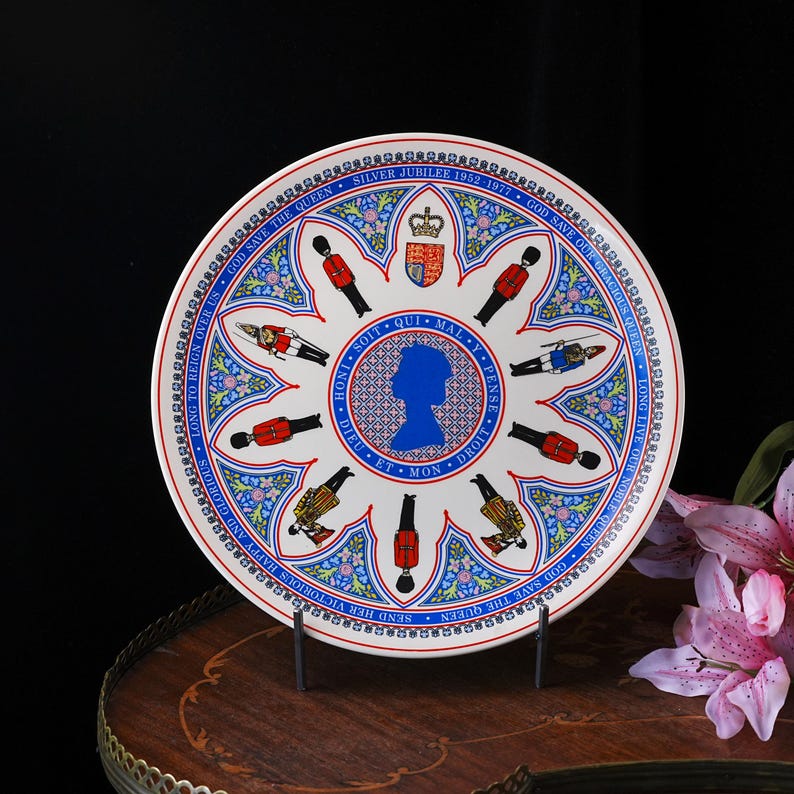 Puede incluir: Un plato conmemorativo de cer&aacute;mica blanca con una silueta azul de la reina Isabel II en el centro, rodeado de ilustraciones de guardias reales, un escudo de armas y motivos florales. El texto dice Silver Jubilee 1952-1977.
