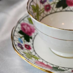 Op de afbeelding: Een witte theekop en schotel met een bloemenpatroon en gouden rand. Het ontwerp toont roze, paarse en blauwe bloemen. De kop en schotel staan tegen een witte achtergrond, met een delicate, patroonrand.