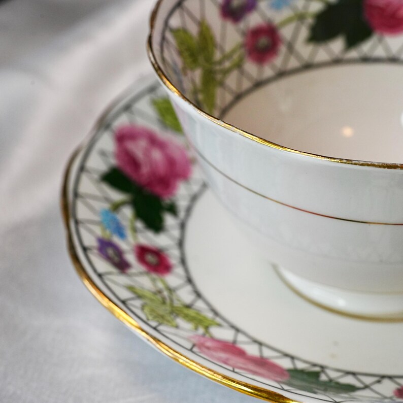 Op de afbeelding: Een witte theekop en schotel met een bloemenpatroon en gouden rand. Het ontwerp toont roze, paarse en blauwe bloemen. De kop en schotel staan tegen een witte achtergrond, met een delicate, patroonrand.