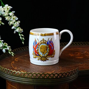May include: A white ceramic mug with a handle, decorated with a royal portrait, flags, and the words "Silver Jubilee" and the dates 1910 and 1935. The mug is displayed on a dark wooden table with a decorative edge.