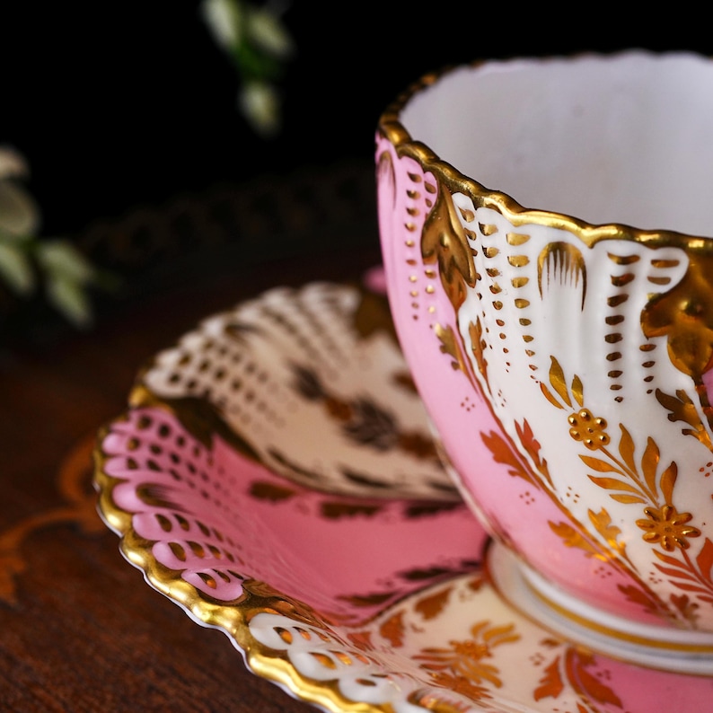May include: A pink and white teacup and saucer set with gold accents. The teacup has a scalloped rim and is decorated with gold floral patterns. The saucer mirrors the teacup's design, creating a cohesive and elegant look.