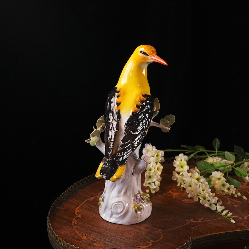 May include: A ceramic bird figurine with yellow and black plumage, perched on a white tree branch. The bird has an orange beak and is set against a dark background. The base of the figurine is white with floral accents.