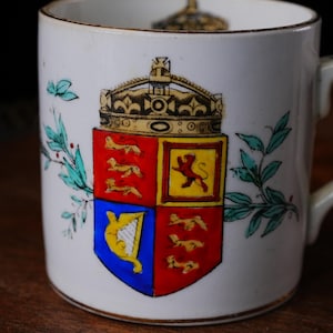 May include: White ceramic mug featuring a colorful coat of arms design with a crown. The crest includes red, yellow, and blue sections with heraldic symbols. Green and blue floral accents are on the side of the mug. The mug has a gold rim.