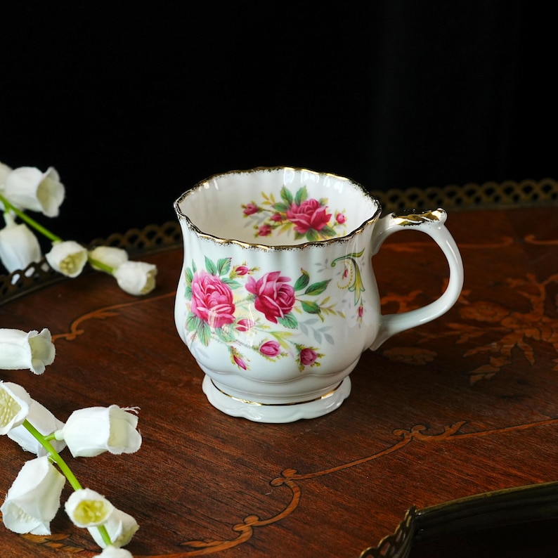Peut inclure: Une tasse &agrave; th&eacute; en porcelaine blanche avec un bord festonn&eacute; dor&eacute; et un motif floral compos&eacute; de roses roses et de feuilles vertes. La tasse pr&eacute;sente une anse incurv&eacute;e et un corps cannel&eacute;, avec des motifs de roses visibles sur le bord int&eacute;rieur.