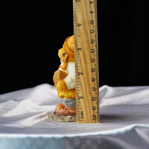 Op de afbeelding: Een klein beeldje van een beer met een hoed en een voetbal. Het beeldje is ongeveer 10 cm hoog. De beer draagt een wit overhemd en gele overalls. Het beeldje staat op een kleine, ronde basis.