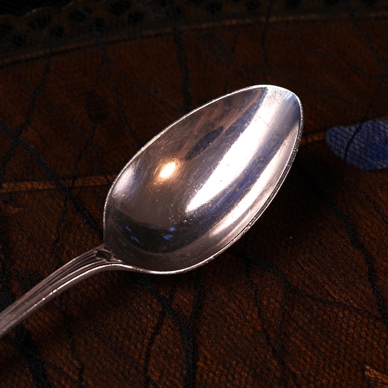Puede incluir: Una cuchara de metal plateado con un cuenco ovalado liso y un mango con sutiles detalles de estr&iacute;as verticales cerca del cuello. La superficie de la cuchara refleja la luz, mostrando un brillo intenso en todo su acabado met&aacute;lico.