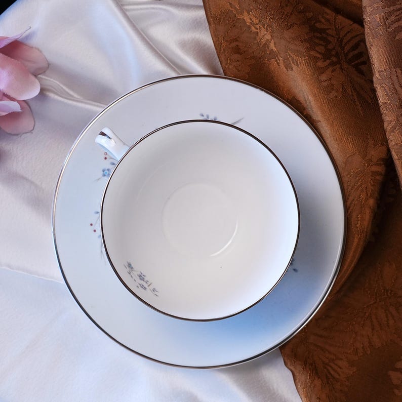 Pode incluir: Conjunto de ch&aacute; branco com ch&aacute;vena e pires, decorado com um delicado padr&atilde;o floral em azul e vermelho. O conjunto tem uma borda prateada e &eacute; colocado sobre um fundo branco e castanho. A ch&aacute;vena est&aacute; vazia.