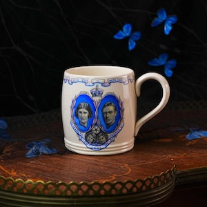 Może przedstawiać: Kremowy ceramiczny kubek z niebiesko-białym wzorem upamiętniającym koronację Elżbiety i Jerzego VI w 1937 roku. Kubek przedstawia portrety pary królewskiej, dwie mniejsze twarze, koronę oraz napis Dieu et mon droit.