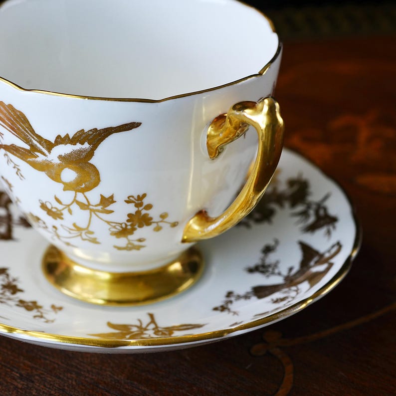 Op de afbeelding: Witte theekop en schotel met gouden vogel- en bloemmotieven. De kop heeft een gouden oor en rand, en de schotel heeft een gouden rand. De set is waarschijnlijk van porselein of fijn porselein.