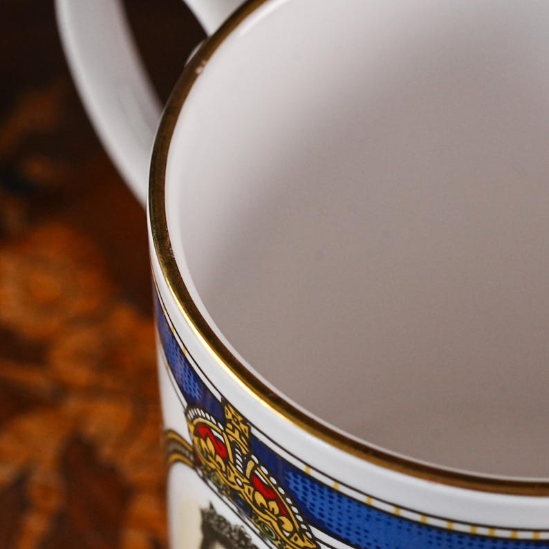 May include: A white ceramic mug featuring a gold-trimmed rim and a blue decorative band. The exterior displays a detailed graphic of a crown in red, yellow, and black colours. The mug has a white handle extending from the side of the cylindrical body.