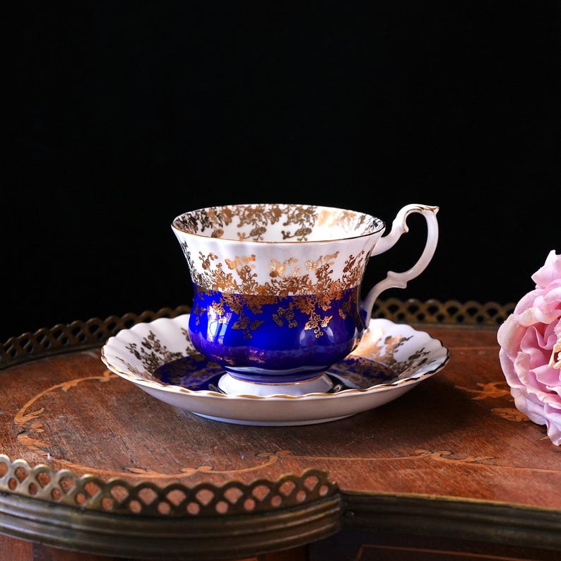 May include: A classic teacup and saucer set. The teacup has a white rim with gold floral patterns, a deep blue body, and a curved handle. The saucer complements the cup's design. The set rests on a wooden tray with a floral pattern.