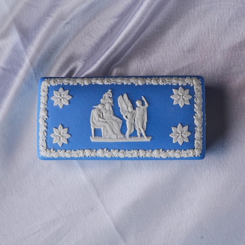 May include: A rectangular blue trinket box with white floral and classical relief details. The box features a central relief scene with figures and angel wings, surrounded by white floral accents.