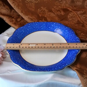 May include: An oval serving platter with a white center and a blue patterned border. The rim has a scalloped edge and a gold trim. The platter measures approximately 12 inches in length.