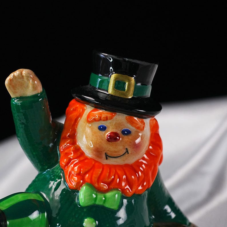May include: A ceramic leprechaun figurine with a green coat, bow tie, and hat. The leprechaun has a red beard, rosy cheeks, and a smiling face. The hat is black with a green band and gold buckle. The figurine is a festive decoration.