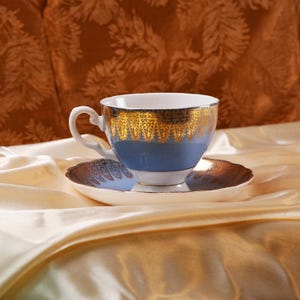 Op de afbeelding: Een blauw en goud theekopje en schotel set. Het theekopje heeft een blauwe body met gouden filigraan rond de rand. De schotel is wit met een gouden rand. De set staat op een cr&egrave;mekleurig satijnen oppervlak.